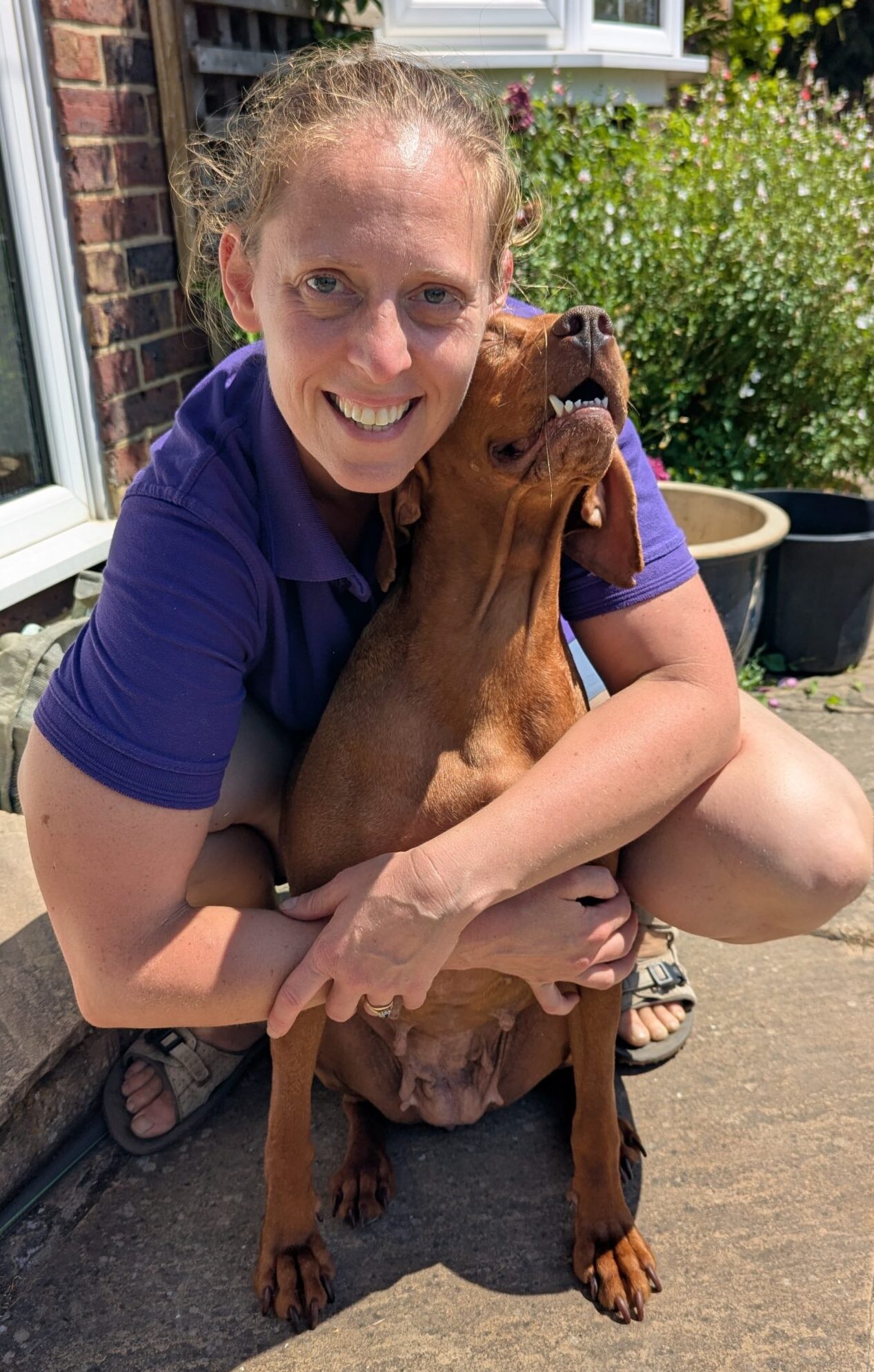 Charlotte and dog - Skylark Vets Skylark Vets Smiling woman in a purple shirt hugging a large brown dog on a sunny front porch with plants nearby.